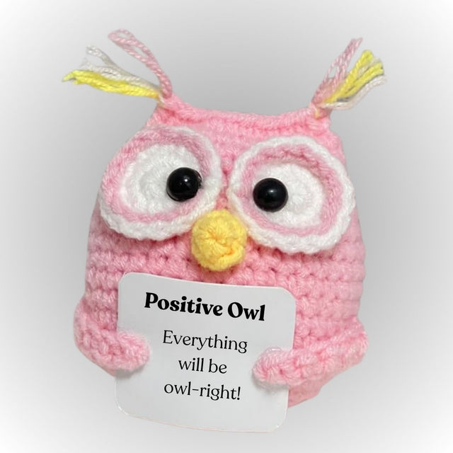 Pink crochet owl with a yellow beak and black eyes, holding a card with text on a white background.